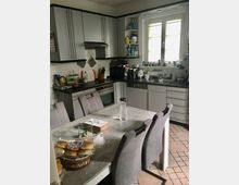 Moderne Küchenansicht mit grauen Hängeschränken, einem Herd mit Dunstabzugshaube, einer Arbeitsfläche mit Küchenutensilien und einem Spülbecken unter einem Fenster mit weißen Rahmen. Im Vordergrund steht ein Esstisch mit grauen Stühlen, auf dem Gebäck, eine Wasserflasche und eine Taschentuchbox liegen.