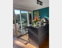 Moderne Küche mit einer Kücheninsel aus dunklem Material, darauf stehen ein Blumenstrauß, eine Espressomaschine und verschiedene Küchenutensilien. Große Glasschiebetüren im Hintergrund geben den Blick auf eine Terrasse mit Holzmöbeln und einen Garten frei.