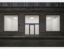 Fassade eines modernen Gebäudes mit großen Schaufenstern, die einen Blick in einen minimalistischen, weißen Innenraum mit Beleuchtung ermöglichen. Rechts ist der Schriftzug „Architektur Galerie Berlin“ sichtbar.