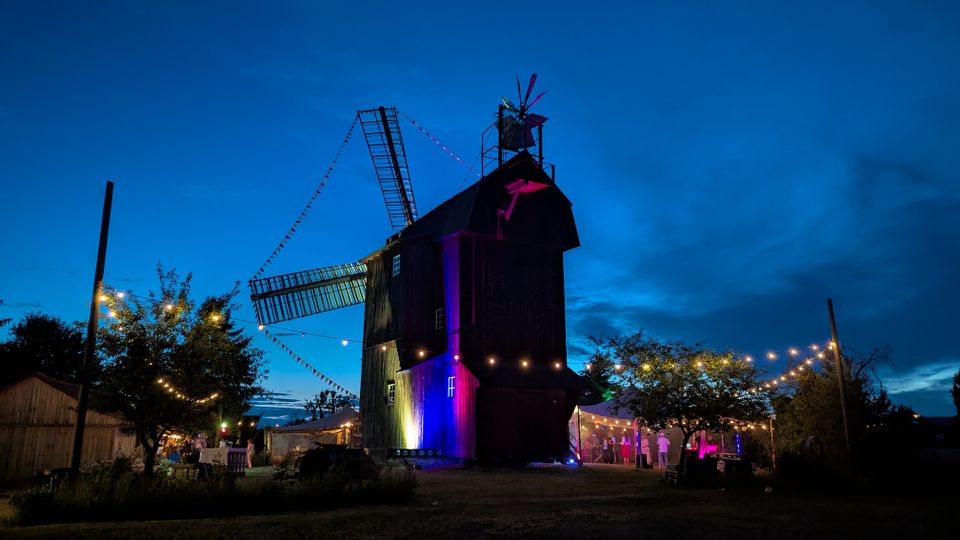 Eine große, hölzerne Windmühle steht in der Abenddämmerung auf einem offenen Gelände, mit langen Flügeln und einem kleinen Aufbau auf dem Dach. Lichterketten und Zelte mit Beleuchtung umgeben die Mühle, Bäume und Nebengebäude rahmen die Szene ein.