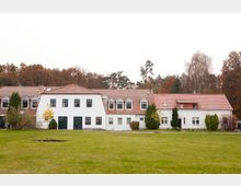 Weiße, einstöckige Gebäude mit roten Ziegeldächern und mehreren Fenstern, umgeben von Bäumen und einer großen grünen Rasenfläche. Im Hintergrund sind herbstlich gefärbte Wälder zu sehen.