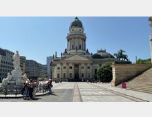 Gebäudeansicht des Deutschen Doms auf dem Gendarmenmarkt in Berlin, mit einer Kuppel und neoklassizistischen Fassadenelementen. Im Vordergrund befinden sich ein Denkmal mit mehreren Figuren, eine Treppe sowie eine Reiterstatue, umgeben von modernem Stadtbau und freiem Himmel.