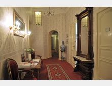 Klassischer Flur mit roten Teppichboden, floralen Tapeten und Wandleuchten. Der Raum ist mit einem kleinen Tisch, roten Polsterstühlen, einem großen Spiegel mit Holzrahmen und einer dekorativen Vase ausgestattet; eine Buntglasfensteröffnung und ein Rundbogen führen in den nächsten Bereich.