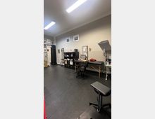 Ein heller, sauberer Raum mit schwarzem Fliesenboden, eingerichtet als Studio oder Arbeitsbereich. Zu sehen sind ein Behandlungstisch, ein Drehstuhl, Regale mit einer Mischung aus Deko und Arbeitsmaterialien sowie Wanddekoration mit gerahmten Bildern.