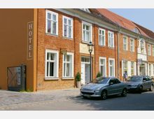 Backsteinfassade eines historischen Gebäudes mit weiß gerahmten Fenstern und einem Schild „Hotel“ an der Seite; davor stehen parkende Autos auf einer gepflasterten Straße, daneben ein schmiedeeiserner Laternenmast.