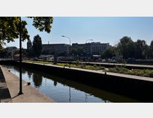 Kanalabschnitt mit gepflastertem Gehweg am Ufer, im Hintergrund Baustellenfahrzeuge und mehrstöckige Gebäude, umgeben von Bäumen. Der Himmel ist wolkenlos, und es sind Laternen entlang des Gehwegs zu sehen.