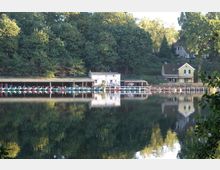 Ein kleiner Bootsanleger an einem ruhigen See, mit mehreren überdachten Bootsständen und einer gelben Villa im Hintergrund, umgeben von dichtem Wald und vereinzelten Häusern. Die Szene spiegelt sich klar im Wasser des Sees wider.
