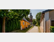 Ein gepflasterter Weg führt zwischen einem gelben Haus und einer Hecke auf der linken Seite sowie einem beige gestrichenen Gebäude auf der rechten Seite zu einer Kirche mit Turm und schwarz spitzem Dach im Hintergrund. Bäume spenden Schatten entlang des Weges.