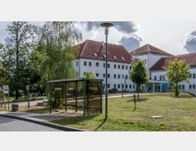 Bushaltestelle mit überdachter Sitzbank im Vordergrund, dahinter ein modernes weißes Gebäude mit rotem Satteldach und runden sowie rechteckigen Fenstern. Die Umgebung besteht aus Grünflächen, Bäumen und vereinzelt platzierten Laternen.