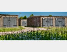 Reihe niedriger Garagengebäude aus rauem Beton mit mehreren grauen Holztoren und einem unbefestigten Weg dazwischen. Im Vordergrund liegt eine grasige Wiese mit Wildblumen, dahinter ist ein blauer Himmel zu sehen.