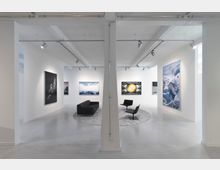 Weißer, moderner Ausstellungsraum mit großformatigen Bildern an den Wänden, minimalistischen schwarzen Möbeln auf einem grauen Teppich und Schienenstrahlern an der Decke zur Beleuchtung der Kunstwerke. Dominierende Architekturmerkmale sind freiliegende weiße Träger und Betonboden.