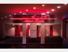 Eingangsbereich eines Kinos mit Treppen, roten Vorhängen und einer Leuchtreklame in roter Neon-Schrift, die den Weg zu weiteren Sälen (2-10) sowie Kassen weist. Die Decke ist mit mehreren runden, eingelassenen Leuchten versehen.