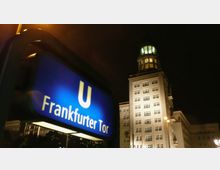 Nachtszene am U-Bahnhof Frankfurter Tor in Berlin, mit einem beleuchteten blauen U-Bahn-Schild im Vordergrund und einem beleuchteten historischen Gebäude mit Kuppel im Hintergrund.