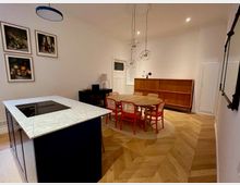 Ein helles Esszimmer mit hölzernem Fischgrätparkett, einer runden Holztischgruppe mit roten Stühlen und einer Kücheninsel mit weißer Marmoroberfläche. An der Wand hängen Stillleben-Gemälde, während eine hölzerne Anrichte und eine moderne Lampe das Interieur abrunden.