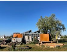 Eine Freifläche mit einem kleinen Gebäude aus Glas und orangen Containern, umgeben von Wiesen und Bäumen. Im Vordergrund sind eine Sandfläche, Holzplattformen und Paletten zu sehen, im Hintergrund eine ländliche Landschaft und weitere kleine Strukturen.