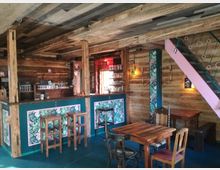Holzinterieur einer kleinen Bar mit rustikaler Einrichtung, bestehend aus Holztischen, Stühlen und einer Theke mit floralen Mustern. Links befindet sich eine Bar mit Gläsern und Regalen, rechts eine farbige Treppe, Wände sind aus Holzplanken mit dezentem Dekor.