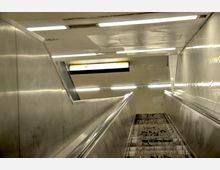 Ansicht einer Rolltreppe in einer U-Bahn-Station mit metallischen Seitenwänden und einer gelb-weißen Anzeigetafel im Hintergrund. Der Raum ist schlicht gestaltet, mit kühler Beleuchtung und einer teilweise schmutzigen Treppe.