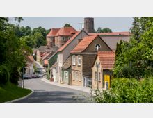 Straße in einem kleinen Dorf mit Reihen von Häusern in unterschiedlichen Farben und roten Ziegeldächern, umgeben von grünen Bäumen. Im Hintergrund ist ein historisches Gebäude mit Turm und rotem Dach sichtbar.