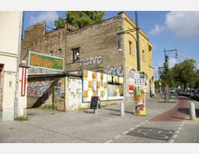 Straßenszene mit alten Gebäuden, darunter eine gelbe Fassade und eine verwitterte Backsteinwand mit Graffiti. Im Vordergrund Werbetafeln, ein Gehweg mit einer Radspur und Straßenlaternen; im Hintergrund Bäume und geparkte Autos.