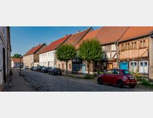 Kopfsteingepflasterte Straße in einer Altstadt mit parkenden Autos, gesäumt von niedrigen Wohnhäusern mit roten Ziegeldächern und teils sichtbarem Fachwerk. Entlang der Häuser stehen gleichmäßig geschnittene Bäume, im Hintergrund verläuft die Straße leicht nach links.
