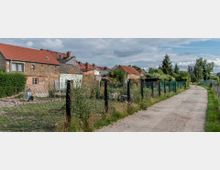 Ländliche Szene mit einer Schotterstraße, die an eingezäunten Gärten und Häusern mit roten Ziegeldächern entlangführt. Die Umgebung ist geprägt von Vegetation, Bäumen und einfachen Wohngebäuden.