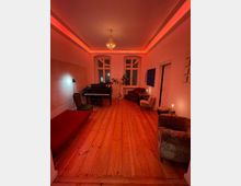 Wohnzimmer mit Holzdielenboden, roter indirekter Beleuchtung und großen Fenstern. Im Raum stehen ein Klavier, verschiedene Sessel, ein Sofa, eine Zimmerpflanze und ein Kronleuchter an der Decke.