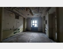 Ein leerer, heruntergekommener Raum mit abblätternder Farbe an Wänden und Decke sowie einem einzelstehenden Fenster, das trübes Licht hineinlässt. Der Raum hat einen gefliesten Boden, beschädigte Belüftungsgitter und eine einfache, veraltete Heizung unter dem Fenster.