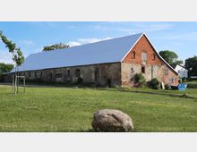 Eine alte, langgestreckte Scheune mit roter Ziegelwand, teils bröckelndem Putz und einem modernen weißen Dach aus Wellblech in einer ländlichen Umgebung mit Rasenfläche und einem großen Stein im Vordergrund. Im Hintergrund sind Bäume und vereinzelte Häuser sichtbar.