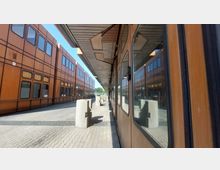 Blick auf einen Bahnsteig mit braunen Gebäuden im Stil moderner Architektur, großen Fenstern und symmetrischem Fassadendesign. Im Vordergrund befindet sich ein abgestellter Zug mit reflektierenden Fensterscheiben, während Betonpfeiler den Gehweg säumen.