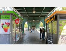 Ein überdachter Gehweg unter einer grünen Stahlkonstruktion einer Brücke oder Hochbahn. Auf beiden Seiten befinden sich Imbissstände, links mit bunten Getränken beworben, rechts für Döner und Kebap. Menschen laufen oder stehen an den Ständen an.