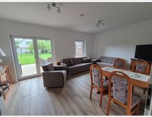 Helles Wohnzimmer mit Essbereich, grauem Bodenbelag und Sitzgruppe aus Sofa und Sessel; nebenan steht ein Esstisch mit gepolsterten Stühlen und ein Fernseher auf einem Lowboard. Große Terrassentüren führen auf eine gepflasterte Terrasse und in einen grünen Garten.