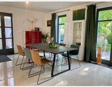 Essbereich mit modernem, rechteckigem Tisch und sechs Stühlen im zeitgenössischen Stil. Die Umgebung umfasst bodentiefe Fenster mit grünen Vorhängen, eine rote Kommode, eine weiße Vitrine und dekorative Elemente wie Kunstwerke und Pflanzen, mit Blick auf einen Garten im Hintergrund.