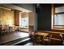 Ein gemütlicher Innenraum eines Cafés oder Restaurants mit Holzmöbeln; links im Bild befinden sich Tische und Stühle vor einer goldgelben Wand mit hängenden Glühbirnen. Rechts sind ein kleines Holzfensterbrett, ein hoher Lautsprecher an der Wand und ein Tisch mit vier Stühlen sichtbar.