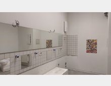 Ein einfacher, leerer Waschraum mit weiß gekachelten Wänden und einem großen Spiegel an der Wand. Einige Wasseranschlüsse ohne Armaturen sowie bunte Poster und ein Handtuchhalter sind sichtbar, der Raum wirkt renovierungsbedürftig.