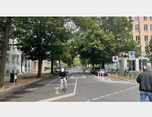 Straßenszene in einer urbanen Umgebung mit breiten Fahrradwegen, flankiert von Bäumen und Wohngebäuden. Mehrere Schilder weisen auf eine Fahrradstraße hin, und es sind Fußgänger, Radfahrer und geparkte Autos zu sehen.