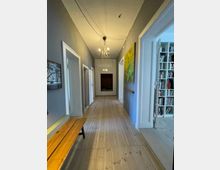 Langer Flur mit hellem Holzboden und grauen Wänden, dekoriert mit Gemälden und einer Hängelampe mit Glasdetails. Links steht eine Holzbank, rechts führt eine Tür in einen Raum mit Bücherregalen.