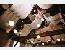 Holzgebälk in einer rustikalen Scheune, geschmückt mit weißen Stoffbahnen an der Decke sowie runden Papierlaternen und Lichterketten, die eine festliche Atmosphäre schaffen. Die Wände bestehen aus sichtbarem Mauerwerk, durchbrochen von einigen Fenstern mit hellem Tageslicht.