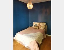 Schlafzimmer mit blau gestrichenen Wänden, einem Holzboden und einem Bett mit rosa Bettwäsche, dekorativen Kissen und einer weißen Decke. An der Decke hängt ein Kronleuchter, und neben dem Bett steht ein kleiner runder Nachttisch mit einer Lampe.