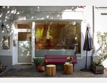 Fassade eines kleinen Cafés oder Ladens mit einer großen Glasscheibe und einer weißen Eingangstür. Vor dem Gebäude stehen eine rote Bank, runde Holzhocker, Blumentöpfe und ein geschlossener Sonnenschirm; auf einem Hocker befinden sich ein Blumenstrauß und eine Teekanne.