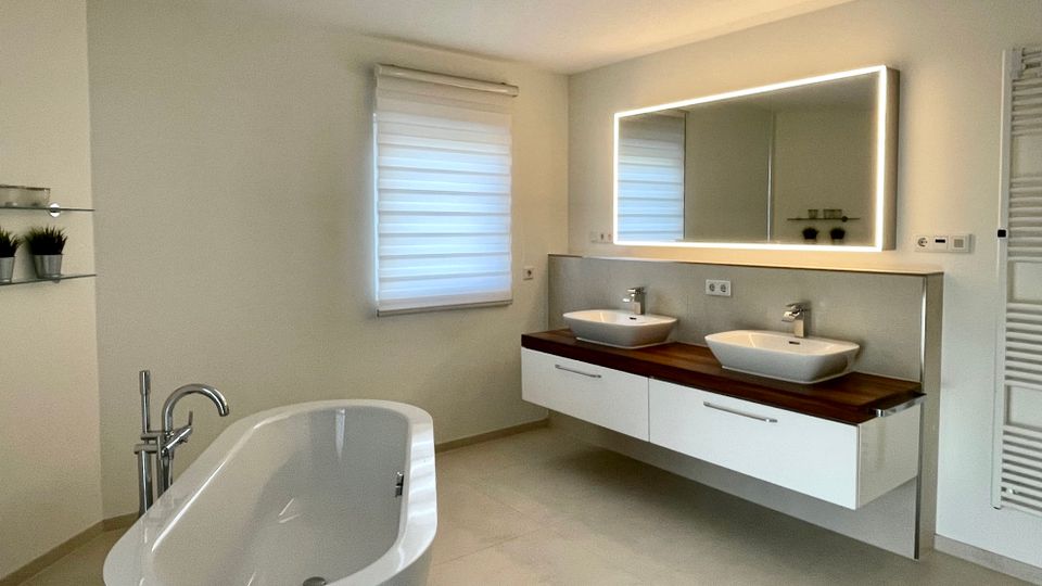 Modernes Badezimmer mit freistehender Badewanne und großem Doppelwaschtisch auf Holzplatte. An der Wand hängt ein breiter, beleuchteter Spiegel über weißen Unterschränken; daneben sind ein Fenster mit Rollo und ein Handtuchheizkörper zu sehen.