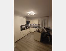 Moderne Einbauküche mit L-förmiger Anordnung, weißen Hochschränken, einer Arbeitsplatte aus dunklem Stein, integrierten Elektrogeräten und Fliesenrückwand. Der Raum hat einen hellen Vinylboden und eine Deckenlampe, rechts steht ein kleines Keyboard neben einem Fenster mit weißen Vorhängen.