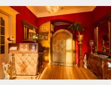 Raum mit opulentem, historischem Stil, geprägt von einer roten Wand, einem goldenen Tor mit detaillierten Verzierungen und antiken Möbeln aus Holz mit Spiegeln. Ein Kristalllüster beleuchtet den Raum, der mit Dekoren wie Figuren, Vasen und Blumenarrangements geschmückt ist.