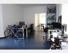 Ein modernes Büro mit weißen Wänden, dunklem Boden und mehreren Schreibtischen, auf denen Computer und Arbeitsmaterialien stehen. Im Vordergrund ist ein Fahrrad zu sehen, im Hintergrund führt ein Durchgang zu einem weiteren Raum mit einem Tisch und Stühlen.