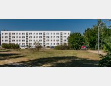 Wohnblock mit vier Stockwerken und weißer Fassade, umgeben von parkenden Autos, Sträuchern und Rasenflächen; rechts im Bild Bäume und eine Straßenlaterne unter klarem blauen Himmel.