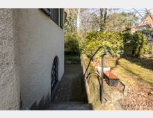 Außentreppenabgang neben einer Hauswand mit Putzstruktur, links ein gitterverkleidetes Kellerfenster, rechts ein schmales Geländer und ein bepflanztes Blumenbeet. Im Hintergrund sind Bäume, Büsche und weitere Gebäude zu erkennen.