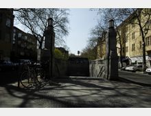 U-Bahn-Eingang am Rüdesheimer Platz in einer städtischen Umgebung, flankiert von zwei steinernen Pfeilern mit Laternen. Im Hintergrund sind Wohngebäude mit gelber Fassade und kahlen Bäumen zu sehen, während Fahrräder im Vordergrund geparkt sind.