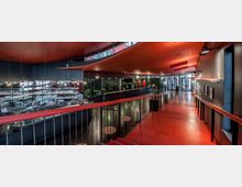 Innenansicht eines modernen Gebäudes mit einem roten Treppenbereich, schwarzen Wänden und Decken sowie einer großen Glasfront im Eingangsbereich. Links befindet sich eine Bar mit glänzenden Oberflächen und reflektierenden Elementen, Sitzmöglichkeiten und beleuchteten Details.