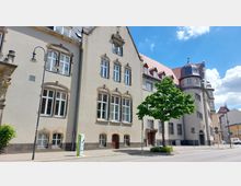 Historisches Gebäude mit heller Fassade, hohen Fenstern mit steinernen Verzierungen und einem Turm mit geschwungenem Dach. Im Vordergrund ein Baum, eine Straßenecke und eine Laterne.