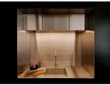 Moderne Küchenzeile aus gebürstetem Edelstahl mit integrierter Spüle und schlichtem Wasserhahn, eingerahmt von hohen Seitenwänden und Oberschränken. Unterbauleuchten beleuchten die Arbeitsfläche, auf der ein Schneidebrett mit Gemüse und Kräutern liegt.