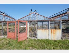 Mehrere verlassene Gewächshäuser mit Glasdächern und rostenden Metallrahmen; im Vordergrund steht eine geöffnete, rote Tür, umgeben von Grasflächen, während der Innenraum der Gewächshäuser leer und bewachsen ist.